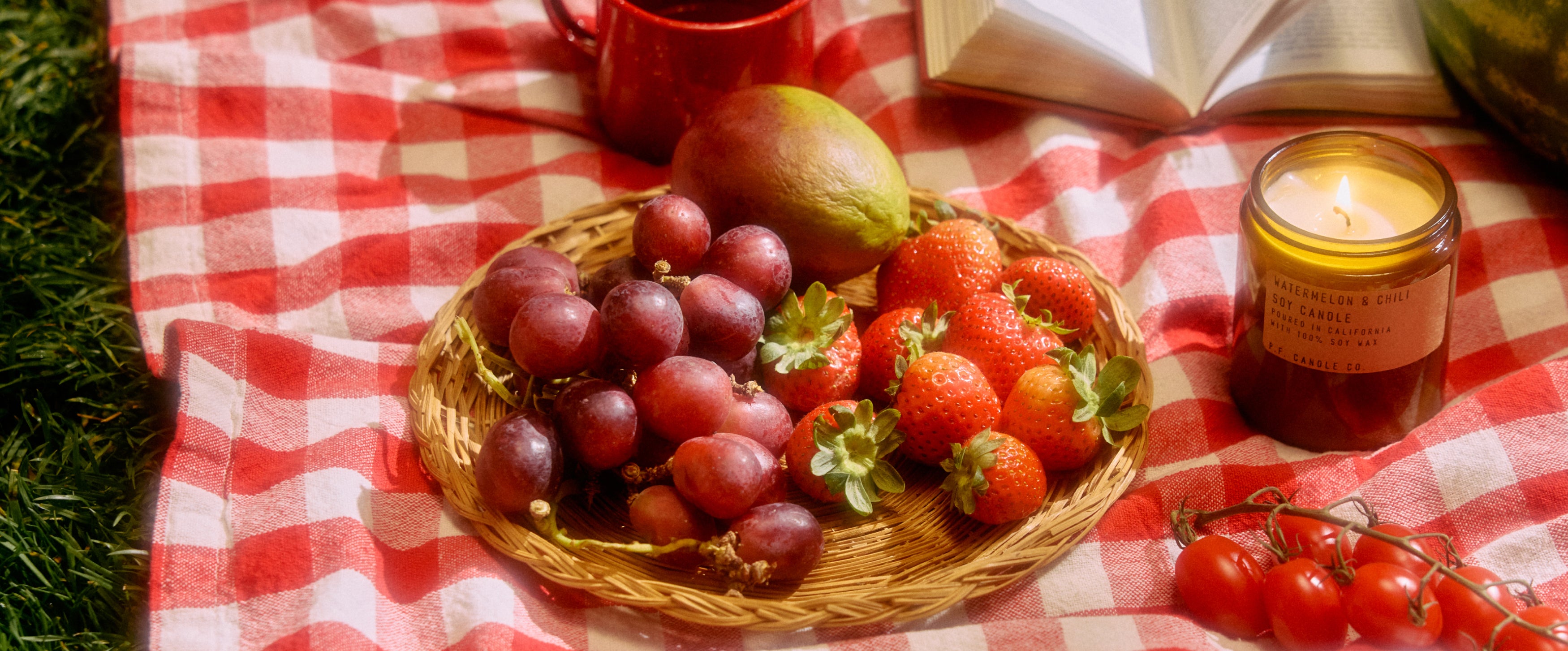 A desktop banner image featuring watermelon & chili candle sitting lit on a picnic blanket with a basket full of  grapes, strawberries and a mango.